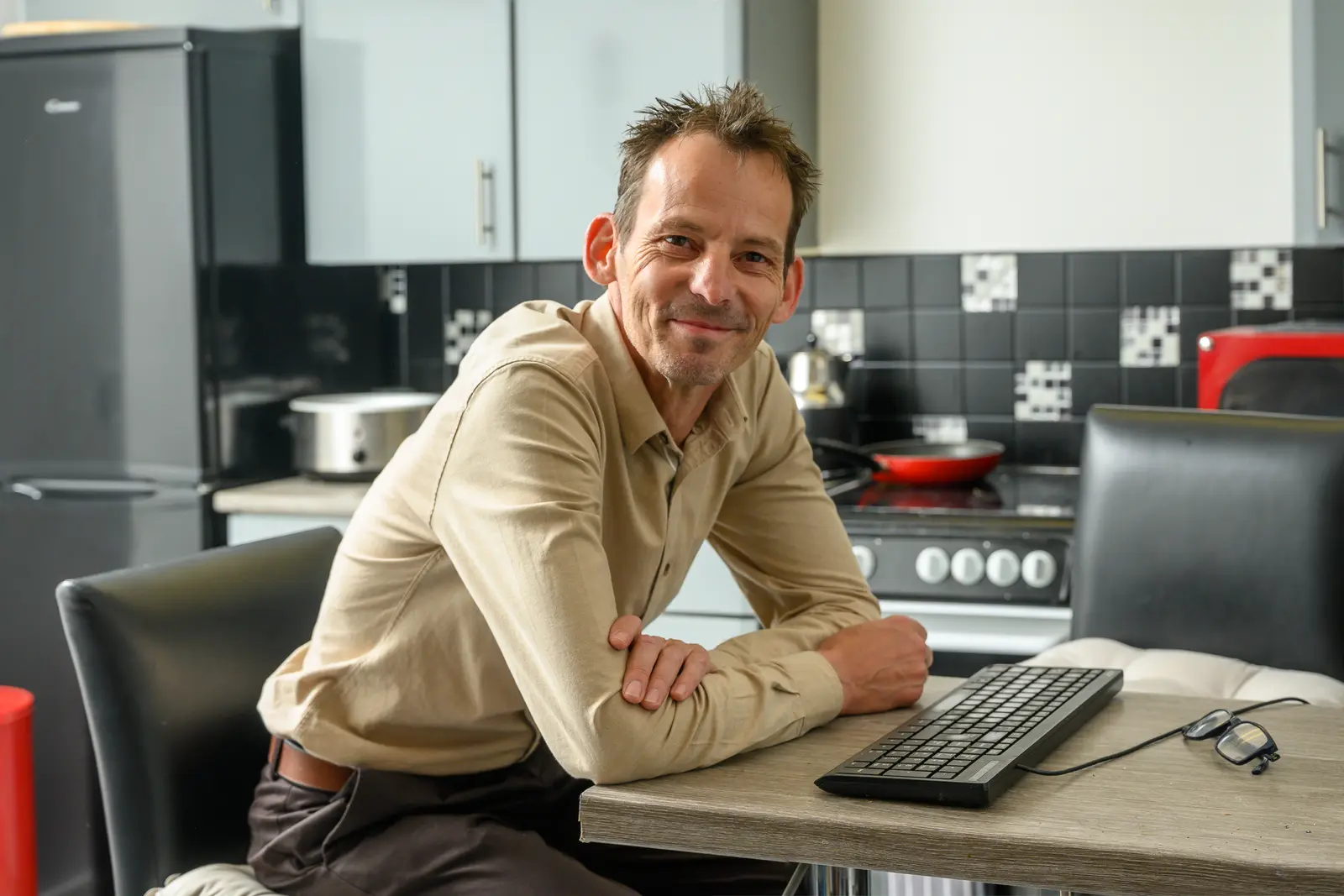 A smiling man sitting at a kitchen table with a computer keyboard
