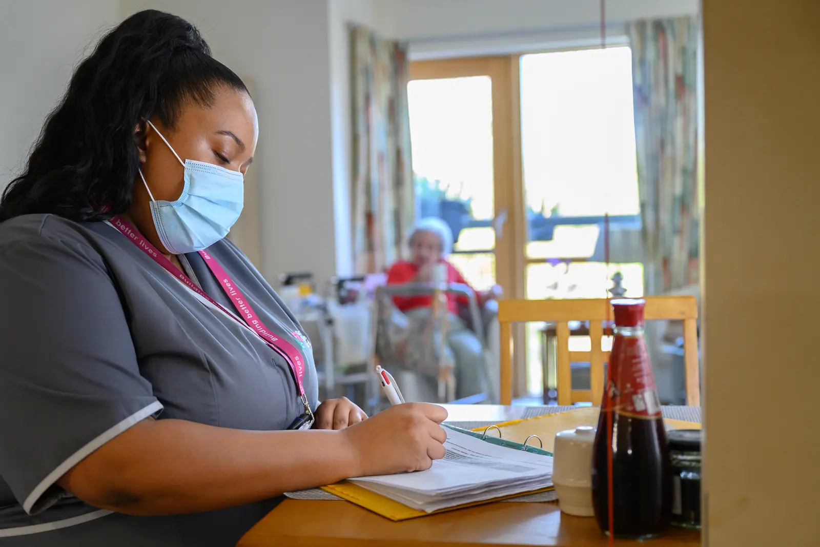 A seated GreenSquareAccord colleague is writing notes while a resident sits behind her