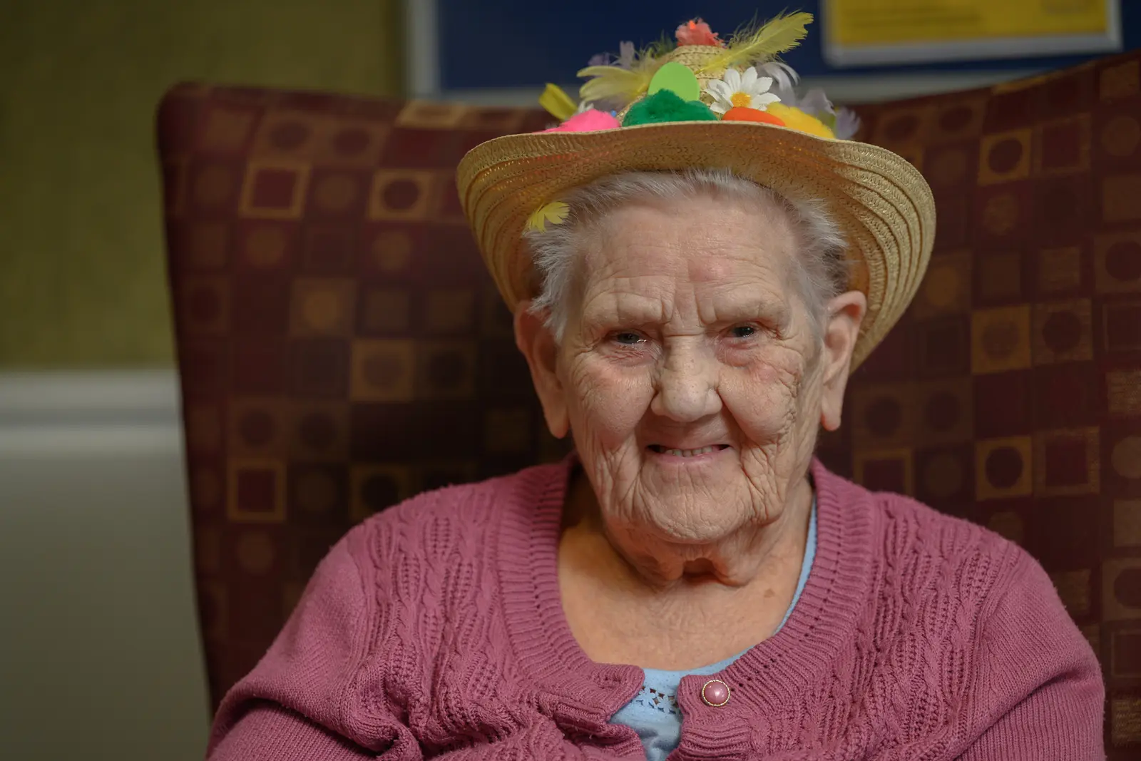 An older lady sitting in an armchair and wearing a straw hat decorated with flowers and feathers