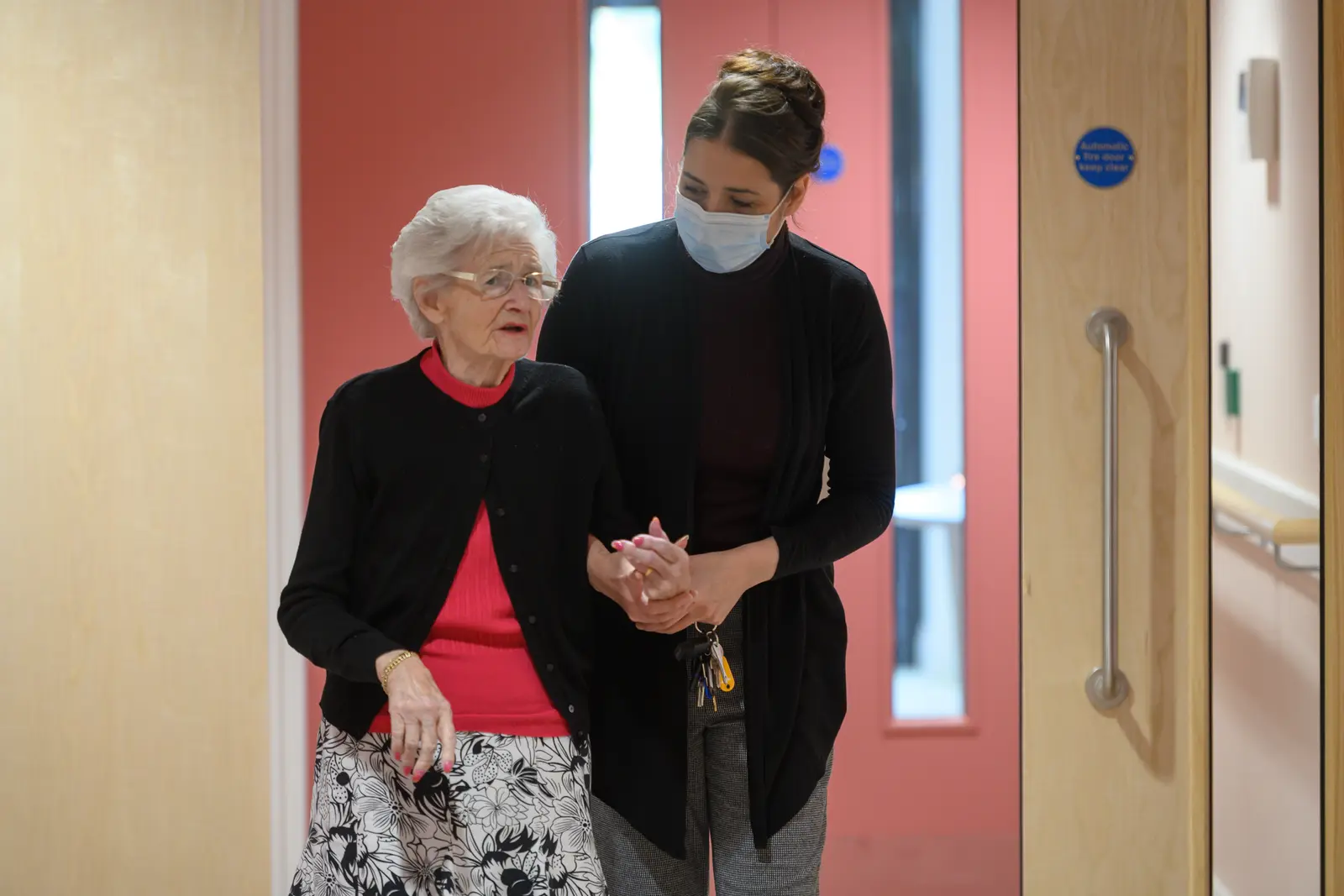 An elderly woman with gray hair walks slowly down a well-lit corridor, supported by her daughter, who gently holds her arm for stability