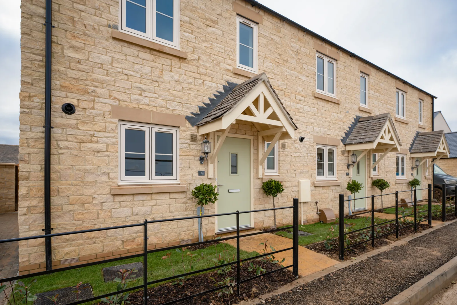 Three traditional Cotswold stone houses with light green doors, each adorned with hanging flower baskets beneath their door canopies.