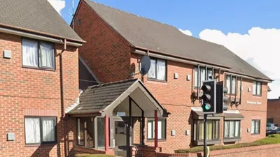 Exterior view of Catherines Court, a two-storey red brick building with a pitched roof and multiple rectangular windows with brown frames. The entrance is covered by a small gabled canopy supported by red columns, leading to a glass doorway. A brick wall runs along the front of the property, separating it from the pavement.