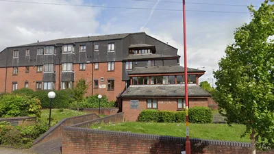 Four‑storey red‑brick residential building at Cherry Tree Court, featuring dark slate‑clad upper levels with dormer-style windows and a stepped roofline. A lower annex with a tiled roof and large windows sits in front, bordered by neatly trimmed bushes and landscaped pathways. The scene includes a brick boundary wall, spherical street lamps, and a tree in the foreground on a bright, partly cloudy day.