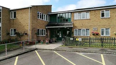 Exterior view of Heberden House, a two-storey brick building with a flat roof and a central entrance covered by a small sloped canopy. The entrance area is framed by green panels and glass windows above the doorway. Several rectangular windows with white frames are visible across both floors. A wooden picket fence runs along the front garden, which is decorated with hanging flower baskets and potted plants.
