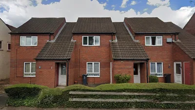 A row of red brick terraced houses with brown tiled pitched roofs. Each house has a front door and rectangular windows with white frames. The roofs feature multiple gables creating a staggered design. A low hedge runs along the front of the properties, with a wooden rail above it.