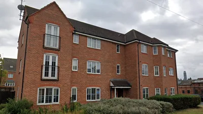 Three-storey red-brick apartment building at Catherines Court with white-framed windows and small metal Juliet balconies, a pitched tiled roof, and a central covered entrance, bordered by low shrubs and planting.