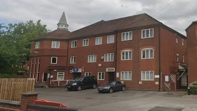 Exterior view of Church House in Walsall, a large three-storey red brick building with a pitched roof and a small white spire on the left corner. The facade features multiple rectangular windows with white frames arranged symmetrically across all floors. Two cars are parked in the paved area in front of the building, which is enclosed by a low brick wall and a wooden fence section. Trees are visible in the background, and the sky is overcast.