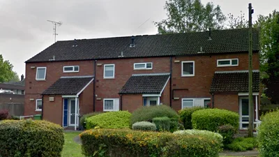 Row of two‑storey red‑brick terraced homes on Collingwood Drive, each with small pitched porch roofs over the front doors, white‑framed windows, and dark tiled roofs. The houses are fronted by neatly maintained shrubs and bushes, with a grassy area in the foreground and mature trees behind the buildings under an overcast sky.