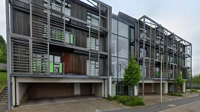 Modern apartment buildings at Daisy Drive with stacked balconies and metal screens, large glass windows, and timber panels, set above covered parking bays along a quiet residential street with young trees and greenery.