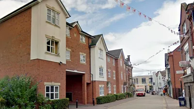 Street view of Eden House, a multi-story red brick residential building with white window frames and detailing. Small shrubs line the base of the building.