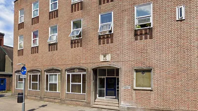 Exterior view of Exchange House, a three-storey building constructed with red brick. The facade features evenly spaced rectangular windows with white frames, some of which are open. The ground floor has larger windows with stone surrounds and a central entrance with a dark blue door set into a recessed doorway. Above the entrance is a decorative stone plaque with an emblem.