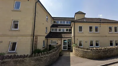 Exterior view of Holton House, a light-coloured stone building with multiple sections and a pitched roof covered in dark tiles. The structure has three storeys, with rows of white-framed windows on each level. The central entrance features a glass door and is flanked by stone walls forming a curved boundary along the pavement.
