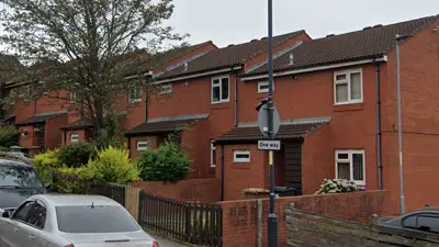 A row of two-storey red brick residential buildings with fenced gardens at the front.