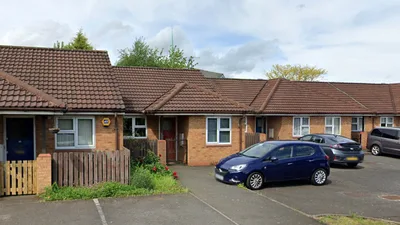 A row of single-storey brick bungalows with brown tiled roofs, each featuring a porch and front door. Three cars are parked outside. The setting is a quiet residential area.
