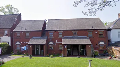 Two‑storey red‑brick residential building at Norton House, featuring a pitched slate roof, arched ground‑floor windows, and two covered entrance porches leading to individual units and an open stairwell. The building faces a well‑kept grassy lawn with scattered potted plants and garden items, framed by neighbouring terraced houses.