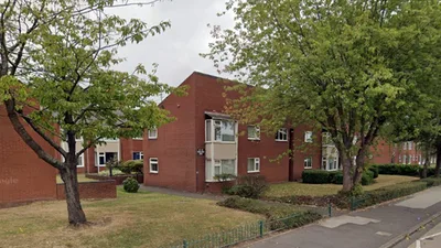 Two‑storey red‑brick residential blocks on Parklands Road in Walsall, arranged around a grassy open area with mature trees. The buildings have white‑framed windows and flat roofs, with one section featuring a small balcony. A narrow pavement runs alongside the road in the foreground, bordered by a low green metal fence and shaded by overhanging trees on a cloudy day.
