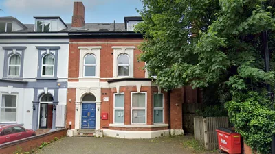 A red brick residential building with two stories, featuring arched windows on the upper floors and a blue front door framed by white decorative trim. The building is part of a row of attached houses and has a paved front area. Trees and greenery partially obscure the right side, where a red Biffa waste bin is positioned near a wooden fence.
