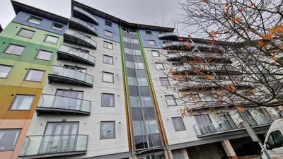 A modern multi-story residential building featuring several balconies and a colorful exterior with panels in green, yellow, and orange. A tree with sparse leaves is visible in the foreground on the right side.