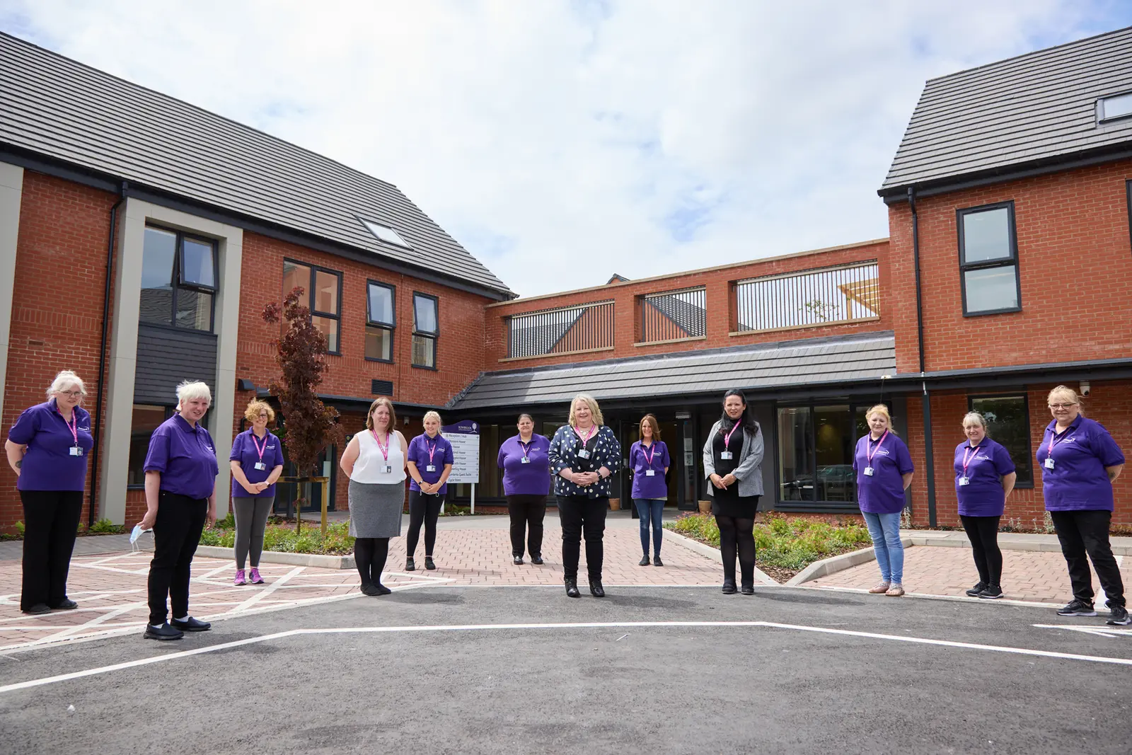 A group of colleagues smiling for a group photo outside a large residential care home on a bright, sunny day.