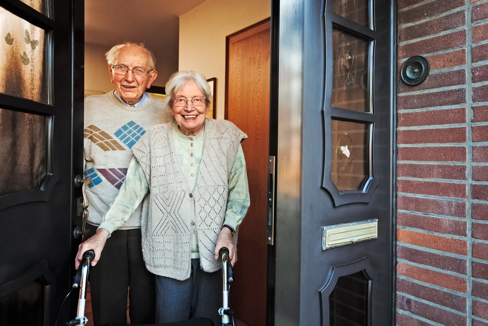 An older lady with a walking frame and her husband stand smiling at their front door